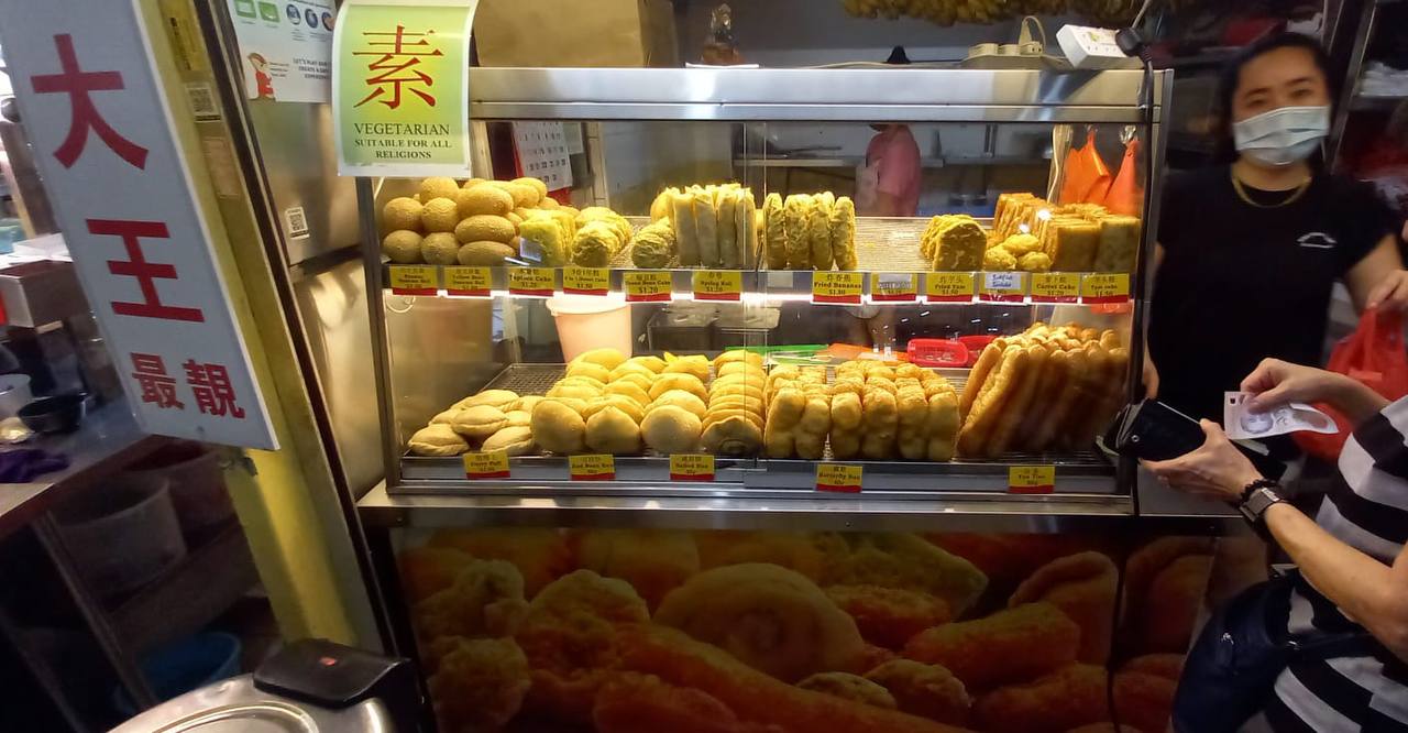the image is a scene of a street food stall in singapore.  the stall is selling a variety of fried asian snacks, such as curry puffs, fried dough sticks, and spring rolls. there are two customers at the stall, one of whom is paying for their purchase. the stall is set up with a glass display case that is filled with the delicious looking treats.