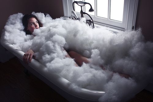 a woman is submerged in a bath filled with fluffy white cotton. the bathtub is in a white bathroom with a window in the background. the cotton is creating a cloudlike effect around the woman, making her look ethereal and serene.