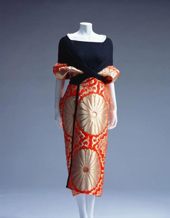 a mannequin wearing a dress. the top part is a black, formfitting top with a square neckline. the skirt is orange with large gold floral patterns and a black band at the waist. the mannequin is standing in front of a gray background. the dress appears to be a unique or designer piece. the dress has a unique design, and the gold and orange combination is eyecatching. the waist part of the dress has a sash made of orange fabric with gold patterns.