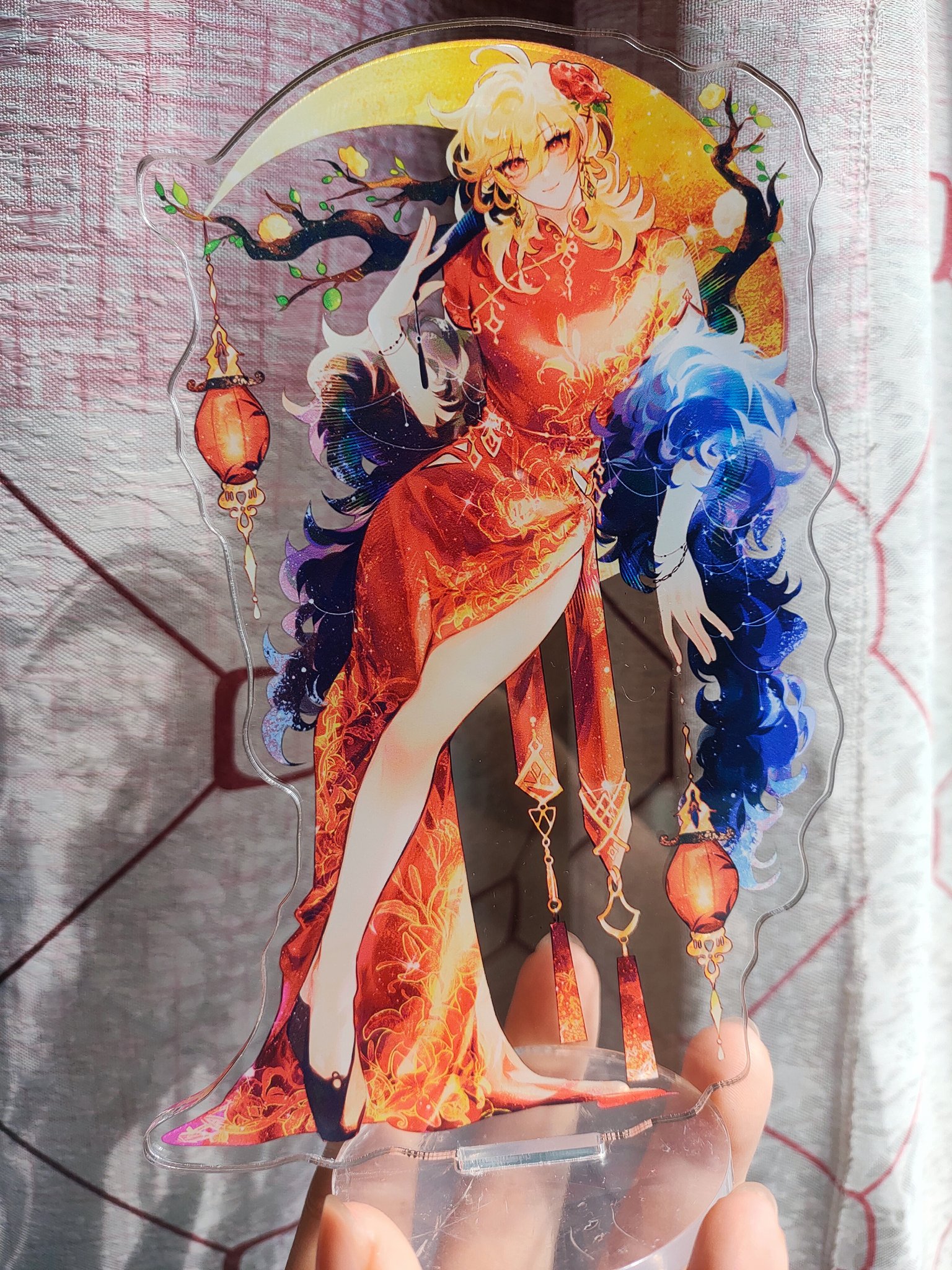 the image shows a hand holding a clear acrylic standup featuring animestyle artwork of a woman. the woman is depicted in a vibrant orange and red qipao (traditional chinese dress), with flowing blonde hair and a dark blue, almost smoky, accessory draped over her shoulder.  she's posed gracefully, with one hand raised and the other resting near her hip. the background of the acrylic piece incorporates a crescent moon, branches of a tree with small fruits, and hanging lanterns, creating a mystical and festive atmosphere.  the artwork is detailed, with sparkles and glowing effects adding to its visual appeal. the entire piece is likely a collectible figurine or piece of merchandise.
