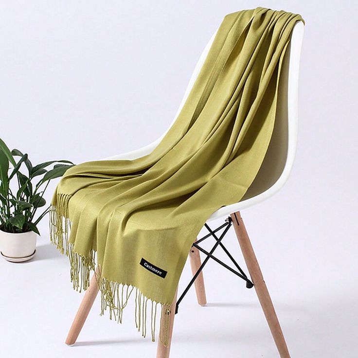 a light green cashmere scarf is draped over a white modern chair with wooden legs and black metal accents. a small green potted plant sits to the left of the chair.