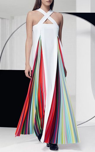 a model wears a long, white halterstyle dress with a colorful, striped skirt that is open in the front. the dress is sleeveless and has a crisscross neckline. the model is standing in front of a geometric background.