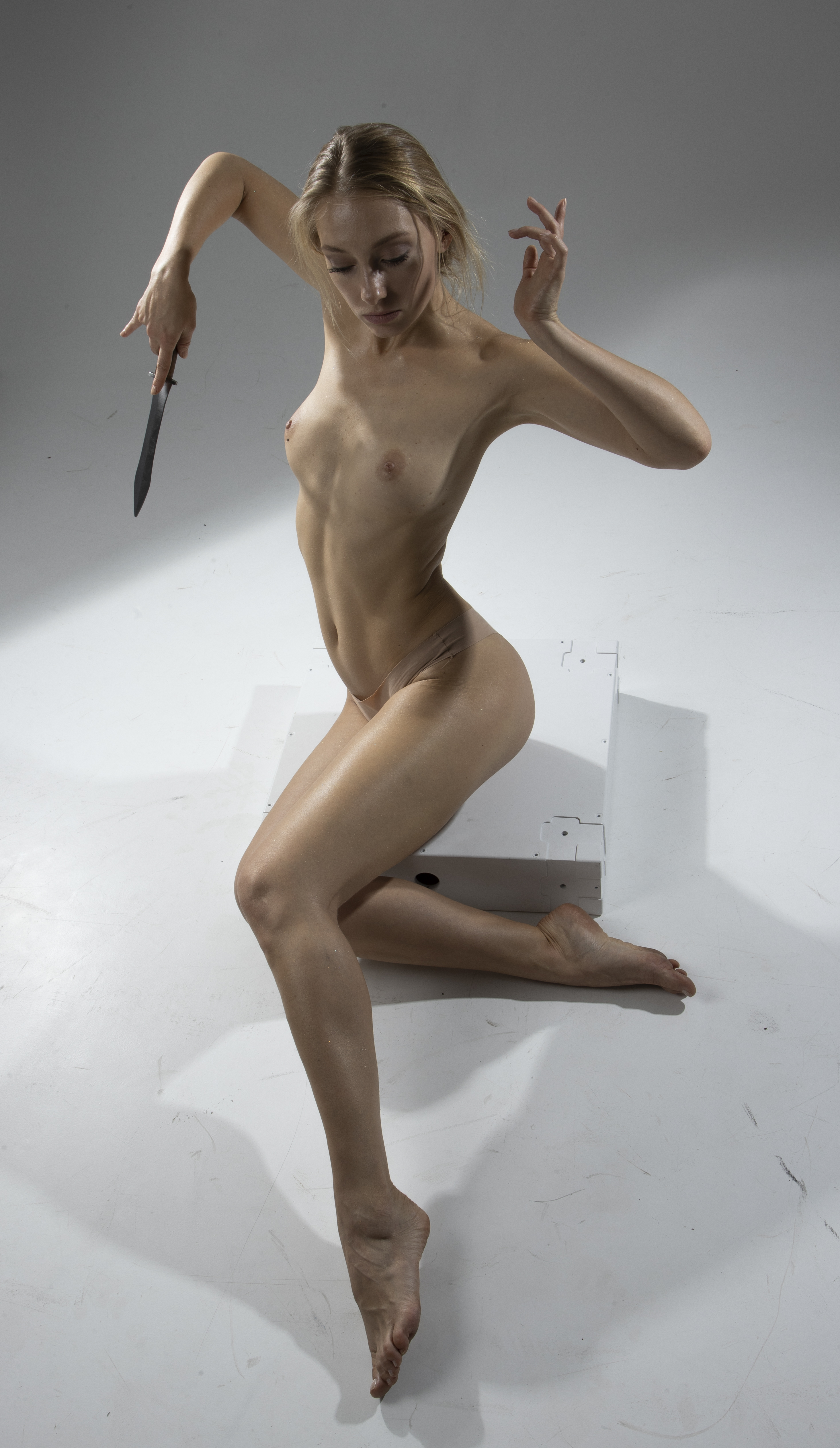 a nude woman with blonde hair is standing in a sensual pose holding a knife against a white background in a studio shot. she has a toned body and fair skin.