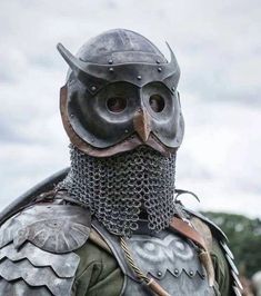 a person stands outdoors, dressed in elaborate armor crafted to resemble an owl.  the helmet is the most striking feature, with a sculpted owl face complete with large eyes and a beak. the wearer's face is obscured by chainmail that hangs from the helmet's lower edge.  the armor continues onto the chest, featuring more metal sculpted with intricate designs. the person appears to be participating in a liveaction roleplaying or cosplay event.