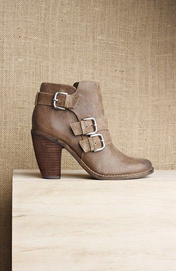 a pair of brown leather ankle boots with buckles are displayed on a light wooden table. the boots are heeled and have a chunky heel. the background is a light brown burlap fabric. the boots are a fashionable choice for a casual or dressedup look.