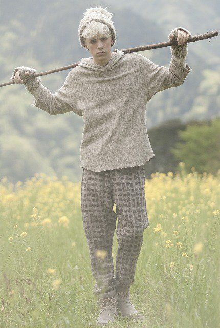 a young man with pale blonde hair, wearing a beige knit beanie, hoodie, and patterned trousers, stands in a field of yellow flowers. he holds a long stick across his shoulders and gazes directly at the camera. the background features rolling green hills, suggesting a serene and natural setting. the lighting is soft and diffused, possibly on an overcast day, lending a gentle and calming atmosphere to the image.