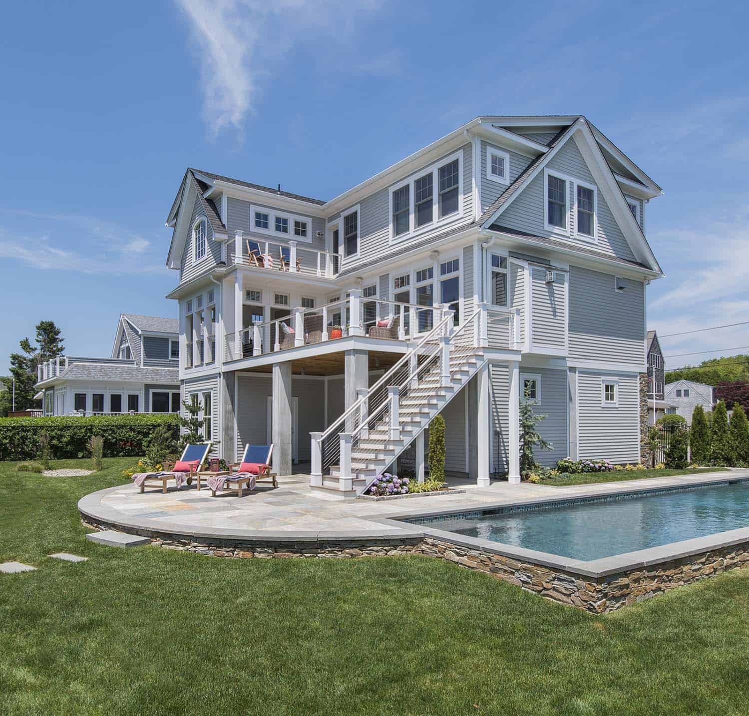 a beautiful light gray threestory home with a backyard pool, patio with lounge chairs, and a manicured lawn. the photo is taken on a sunny day with a clear blue sky.