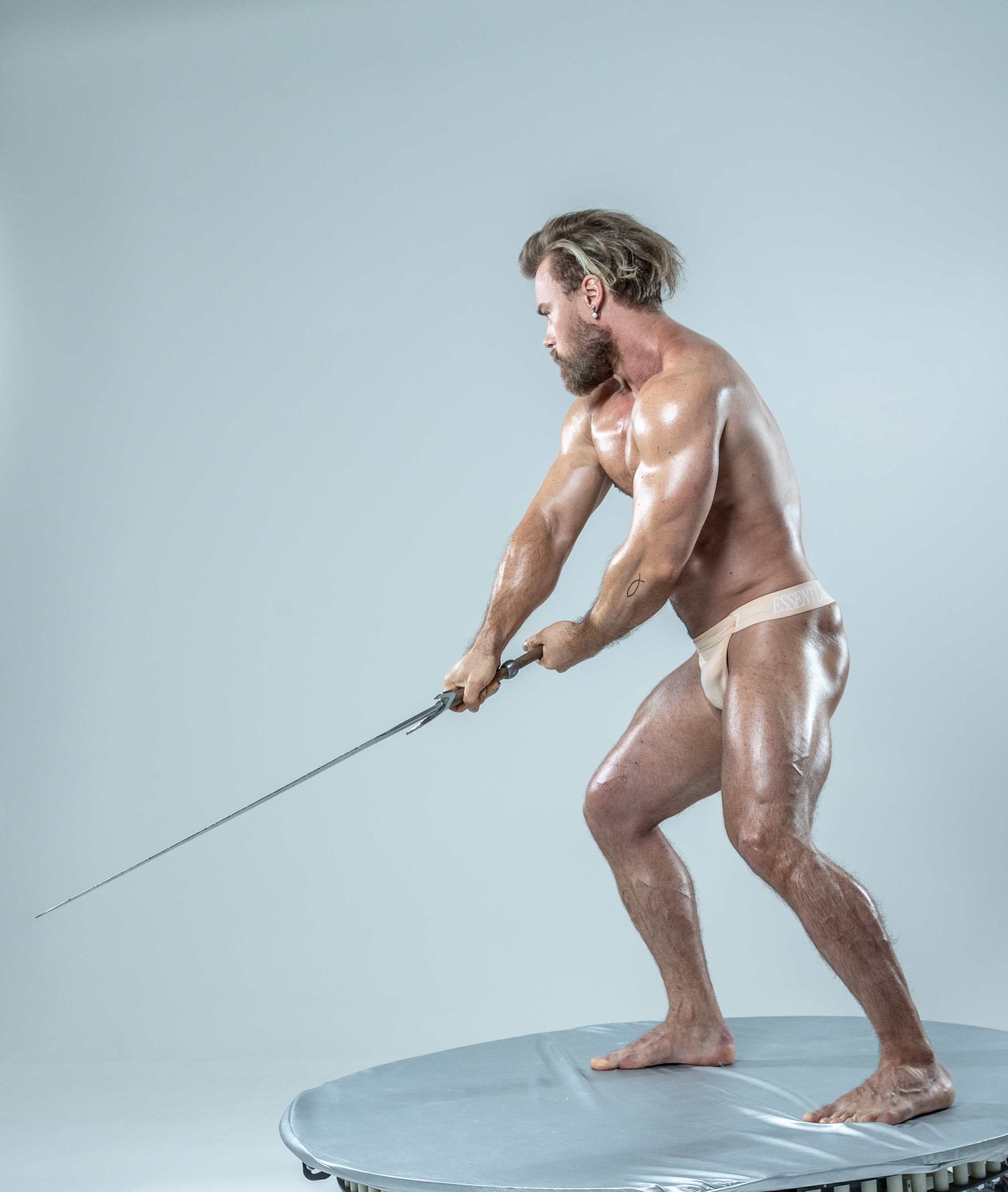 a muscular, bearded man with long blond hair stands on a round platform against a light gray background. he is wearing only a beige loincloth and is captured midswing, powerfully pulling back a long sword. the image evokes a sense of strength, power, and perhaps a historical or mythological theme.  the soft lighting highlights his physique and creates a dramatic atmosphere.