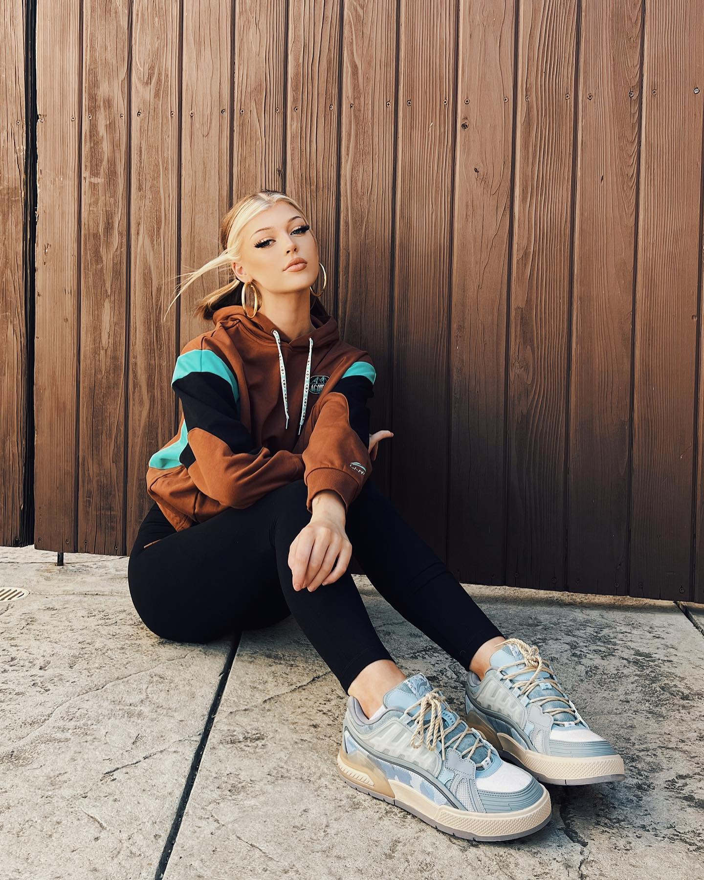 a young woman with blonde hair, hoop earrings, and wearing a brown, black, and blue hoodie with black leggings and sneakers is sitting outdoors against a wooden wall.