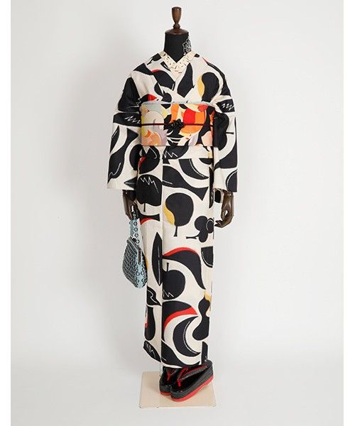 a mannequin is dressed in a striking kimono with a modern, abstract floral design. the kimono features bold black and white shapes, with accents of red, orange, and beige. the mannequin is wearing a vibrant obi with similar geometric patterns, along with traditional geta footwear. a small, light blue handbag is hanging from the mannequin's left hand. the outfit is displayed in a studio setting against a plain white backdrop.