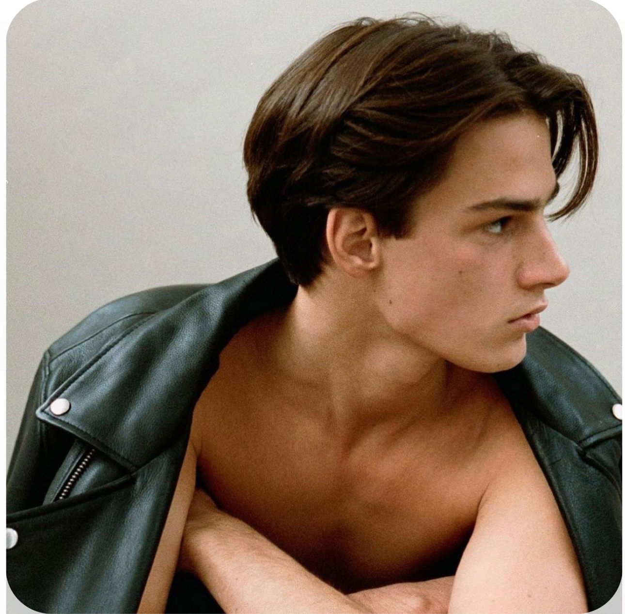 a young man with tousled brown hair wearing a black leather jacket stares seriously to the right of the image. his hair partially obscures his face. only the top half of his torso is visible and he is shirtless beneath the jacket. the background is an offwhite color.