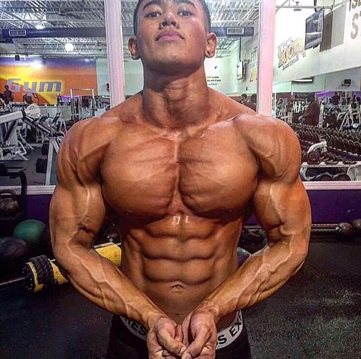 the image shows a muscular asian male bodybuilder posing in a gym. he has highly defined muscles, prominent veins, and a sixpack. his physique is extremely lean and muscular, showcasing the results of intense training. the background shows various gym equipment, including weights and machines, placing him firmly within a fitness context. the overall tone of the picture conveys strength, athleticism, and dedication to bodybuilding.