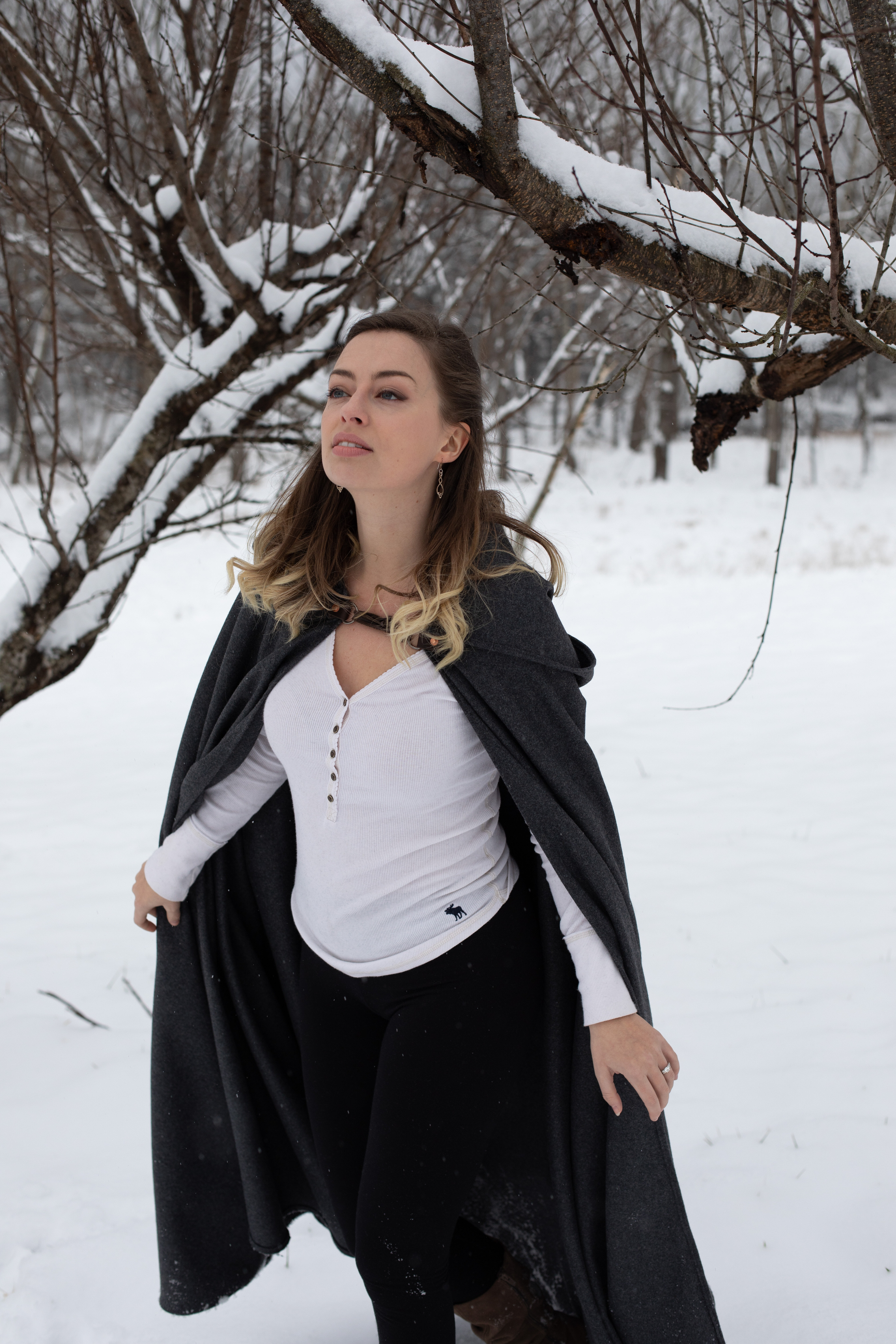 a young woman stands in a snowcovered forest, wearing a long gray cloak over a white shirt and black pants. she is looking upward, her expression serene. the trees surrounding her are bare and covered in snow, creating a peaceful and wintry atmosphere. the image is taken at eye level, providing a natural perspective.