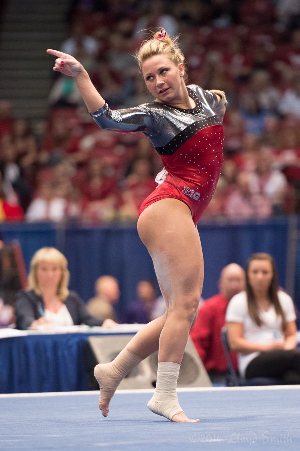 a female gymnast in a red, white, and black leotard with a white ankle brace on each leg is midroutine, performing a tumbling pass on the floor exercise during a competition. she is extending her right arm out, pointing her finger, while extending her left leg straight out behind her. she has a determined look on her face. the crowd is cheering her on in the background. the gymnast is smiling and seems to be enjoying the competition.