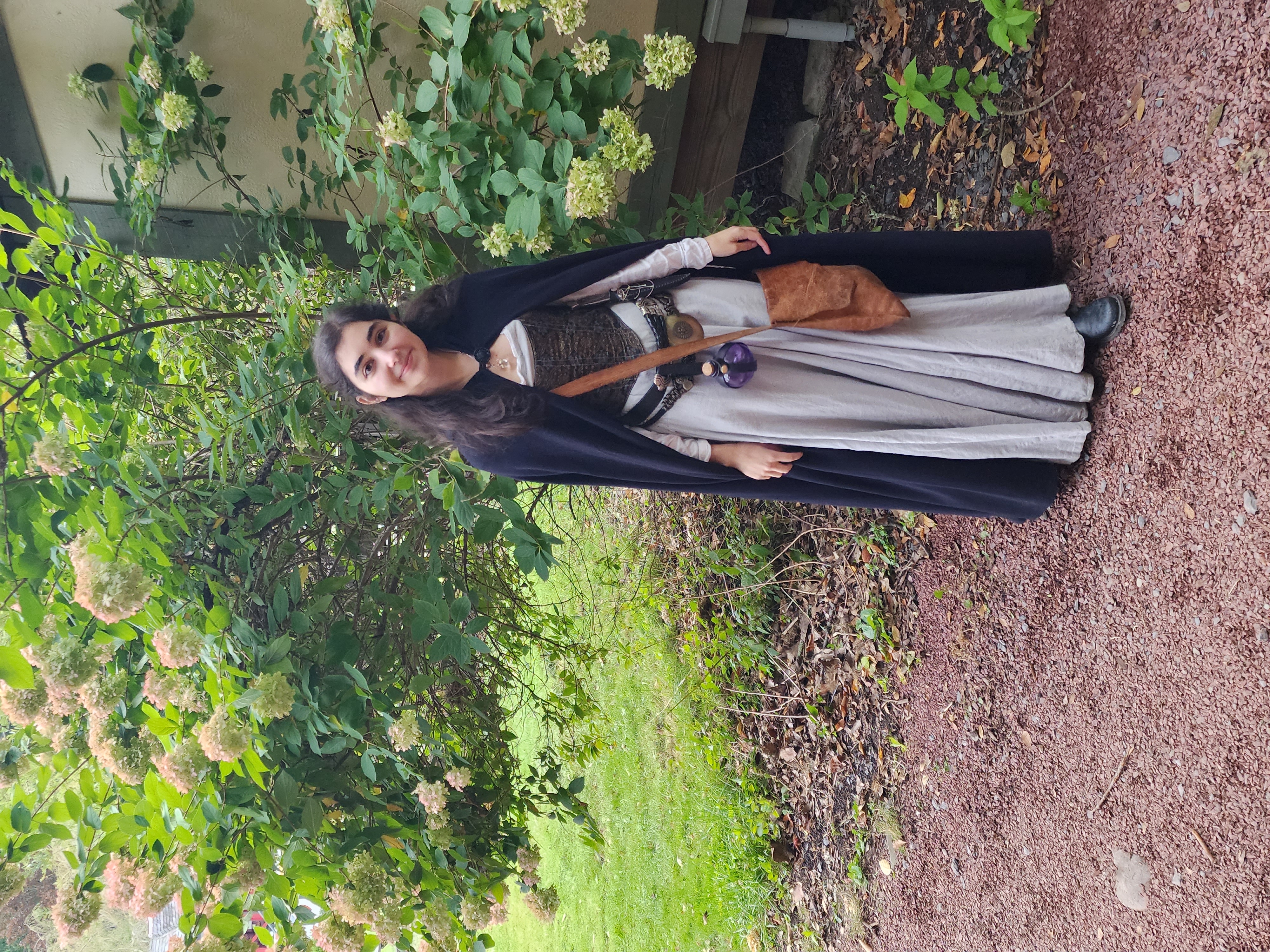 a young woman smiles confidently for the camera, dressed in a medieval or renaissancestyle costume.  she's wearing a long, light gray gown over a brown corset, a black cape, and brown leather boots. a brown leather satchel hangs from her shoulder. the setting is outdoors in a garden with numerous hydrangea bushes in bloom behind her, suggesting a fair or outdoor event.  the background also shows part of a building.  the overall mood is pleasant and whimsical.