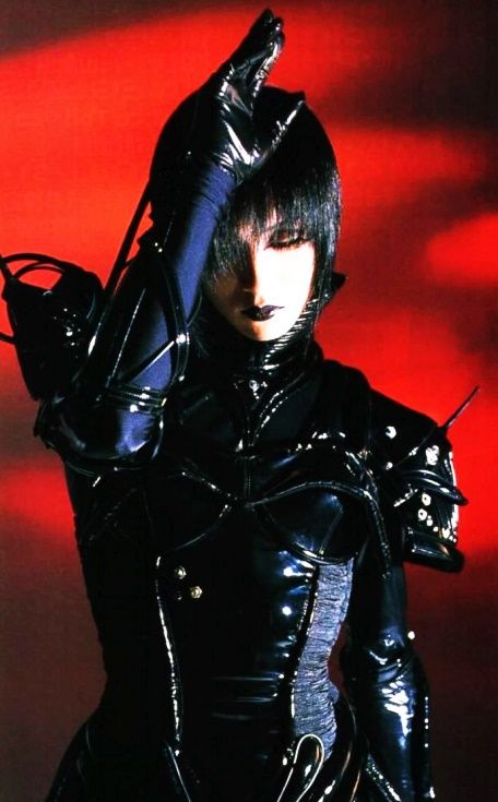 a person in a striking gothic or visual kei style is featured. they are clad in a formfitting black latex outfit with intricate details. the attire includes studded shoulder armor and a high collar. black gloves cover their hands, and they have a dramatic dark makeup look with black lipstick. their hair is styled in a short, dark fringe. the background is a vibrant red, creating a stark contrast with the black outfit. they are making a gesture with their gloved hand near their head. the overall aesthetic is theatrical and edgy.