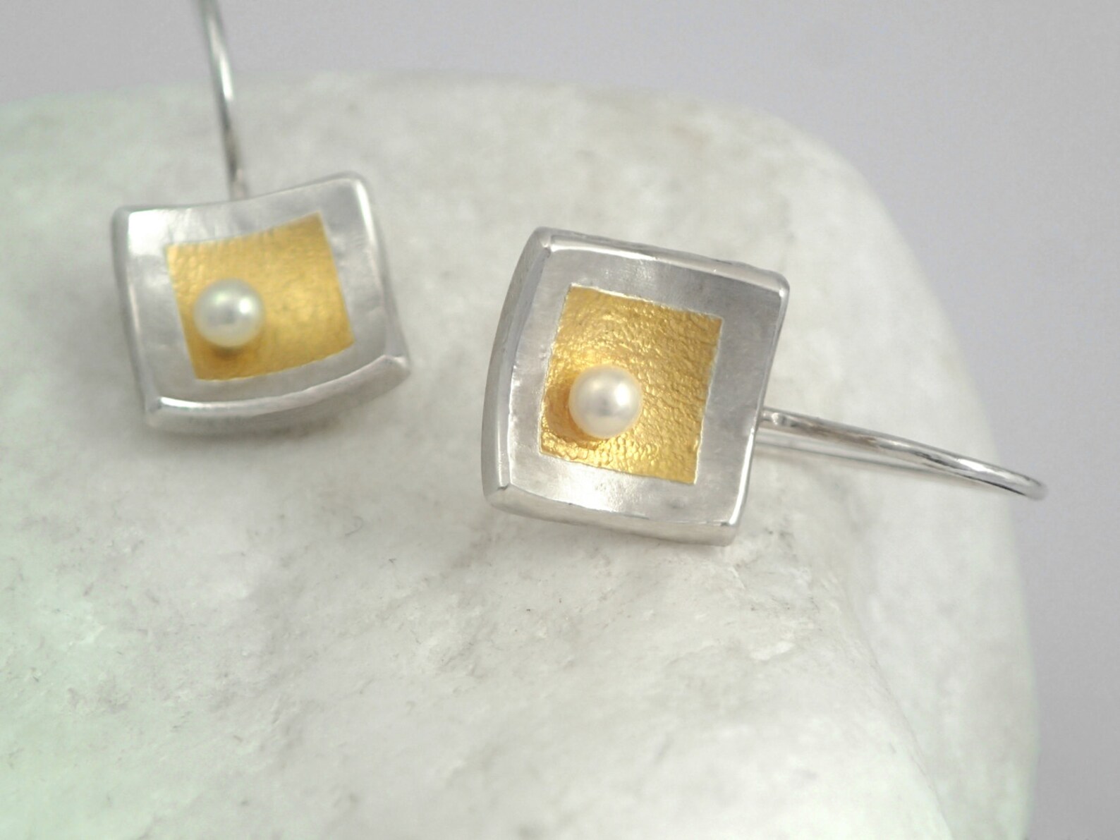this closeup image showcases a pair of intricately crafted earrings. they feature a unique design where a lustrous white pearl is nestled within a square frame. the frame is a striking combination of silver and gold, with the inner square boasting a vibrant gold finish. the earrings rest delicately upon a textured white surface, possibly a piece of natural stone. the image is taken from a top view, highlighting the geometric precision of the earrings' design and the smooth, polished surfaces of the metals. the lighting is bright and even, enhancing the details and creating a sense of elegance.
