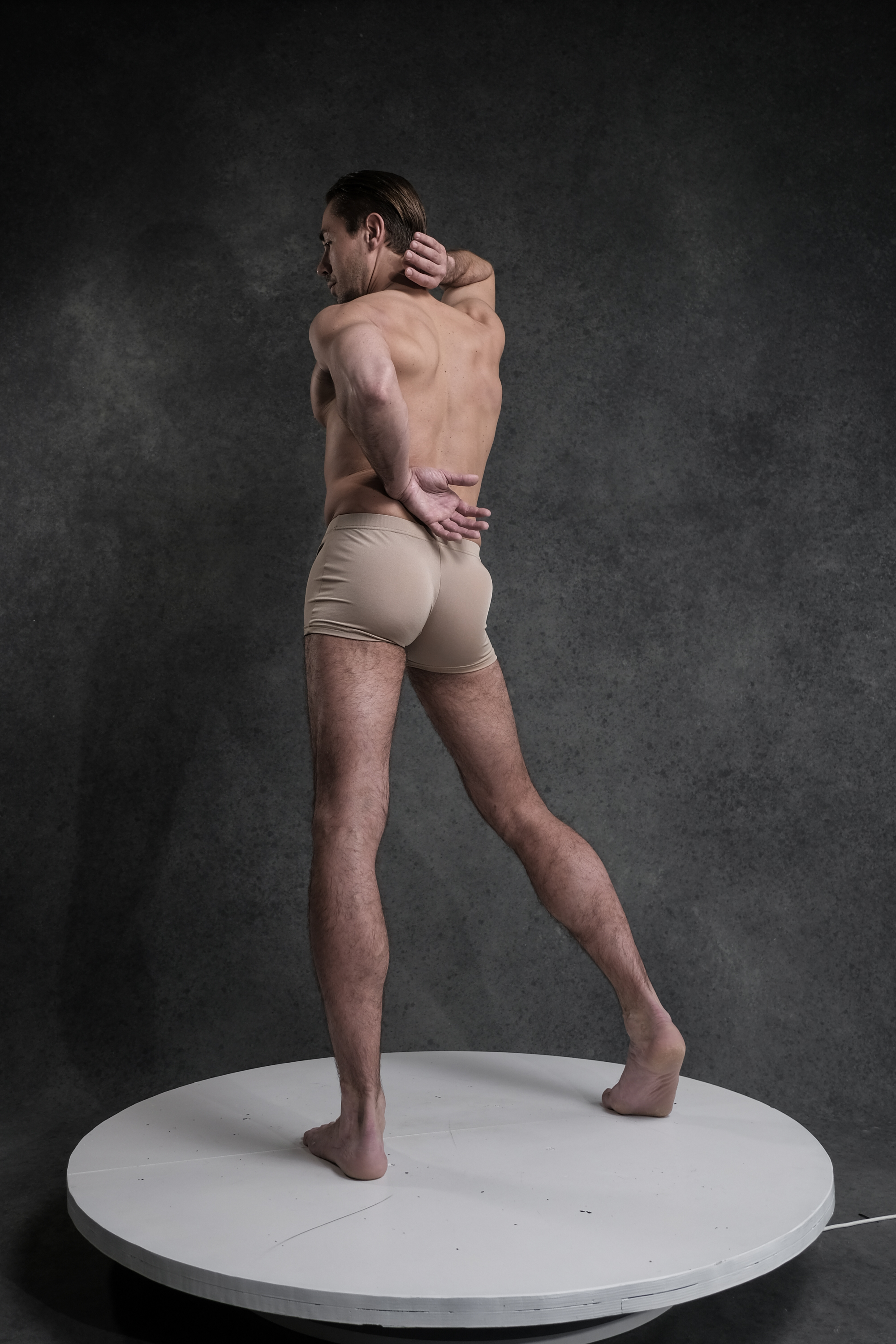shirtless adult male with short hair stands on a white rotating platform against a dark gray backdrop. he is wearing tan underwear and is captured from the rear, with his left hand resting on his lower back and his right hand reaching up to touch the back of his head. his posture suggests a sense of contemplation or perhaps mild discomfort. the studio lighting illuminates his form, highlighting the musculature and contours of his back, legs, and arms.