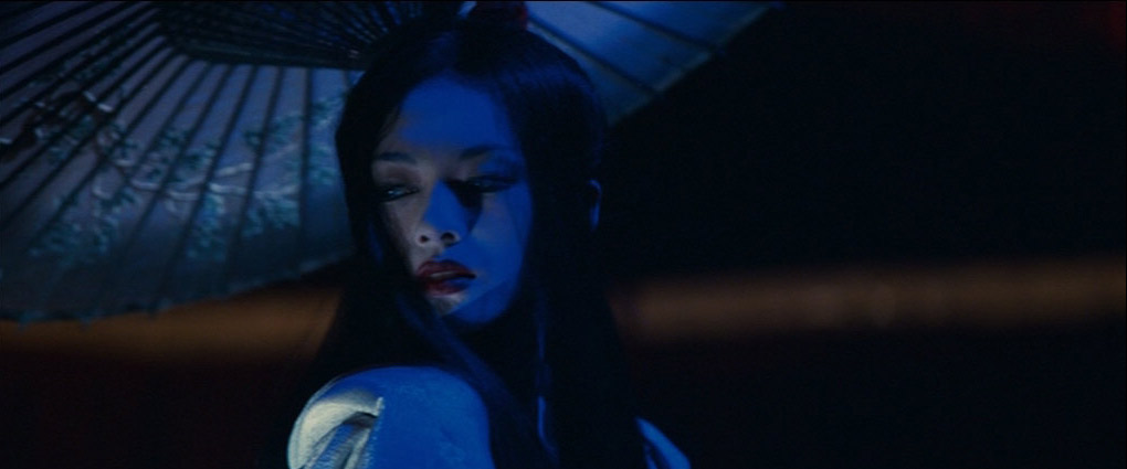 a young asian woman dressed in a dark blue kimono, with long black hair and red lips, looks directly at the camera with a mysterious and alluring expression. the scene is lit with blue lighting, casting dramatic shadows on the woman's face and the background. she's holding a traditional japanese umbrella, and the overall effect is mysterious and sensual.