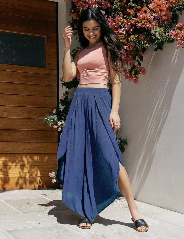 a young woman is standing outside in front of a wooden door and a wall covered with pink flowers. she is wearing a pink crop top and wideleg blue pants with slits on the sides. she is looking down and smiling slightly, with her left hand touching her hair. she is wearing black sandals and has long dark hair. the lighting is bright and natural, creating a cheerful and summery atmosphere.