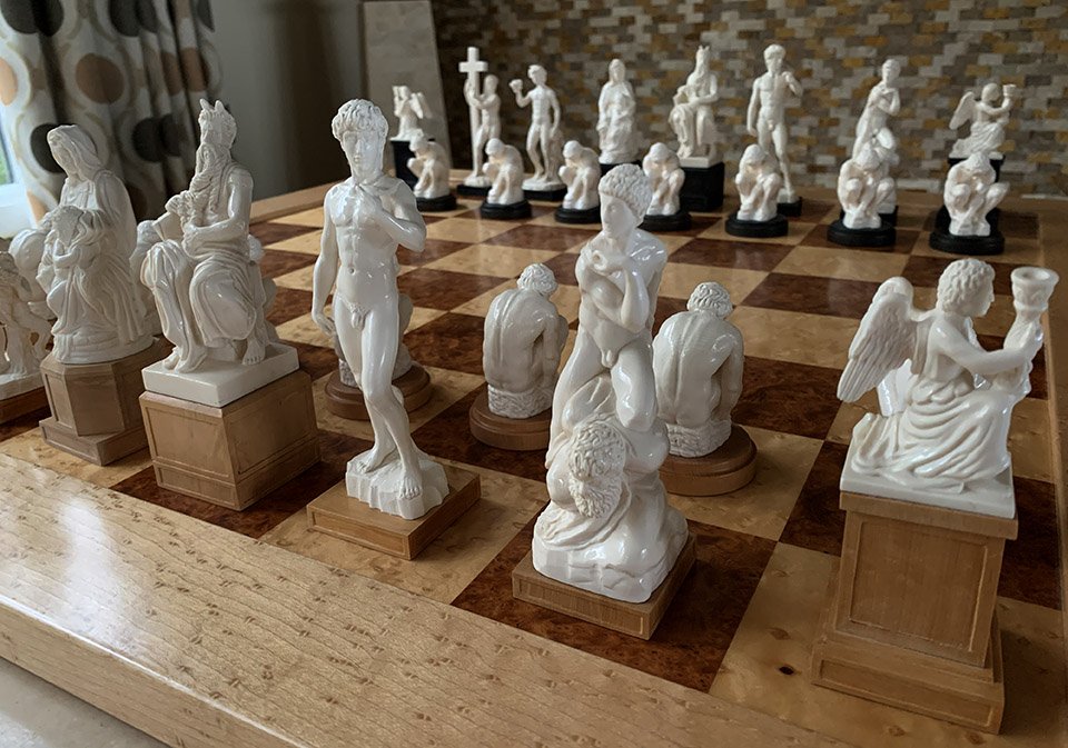 this image showcases a meticulously crafted chess set featuring intricately detailed figurines inspired by classical and mythological themes.  the pieces, possibly made from ivory or a similar material, stand on a luxurious wooden chessboard with alternating light and dark squares.  the setting appears to be indoors with ample natural light highlighting the craftsmanship of the set. the perspective is slightly elevated, offering a clear view of the layout of the chessboard and the positioning of the sculpted pieces.
