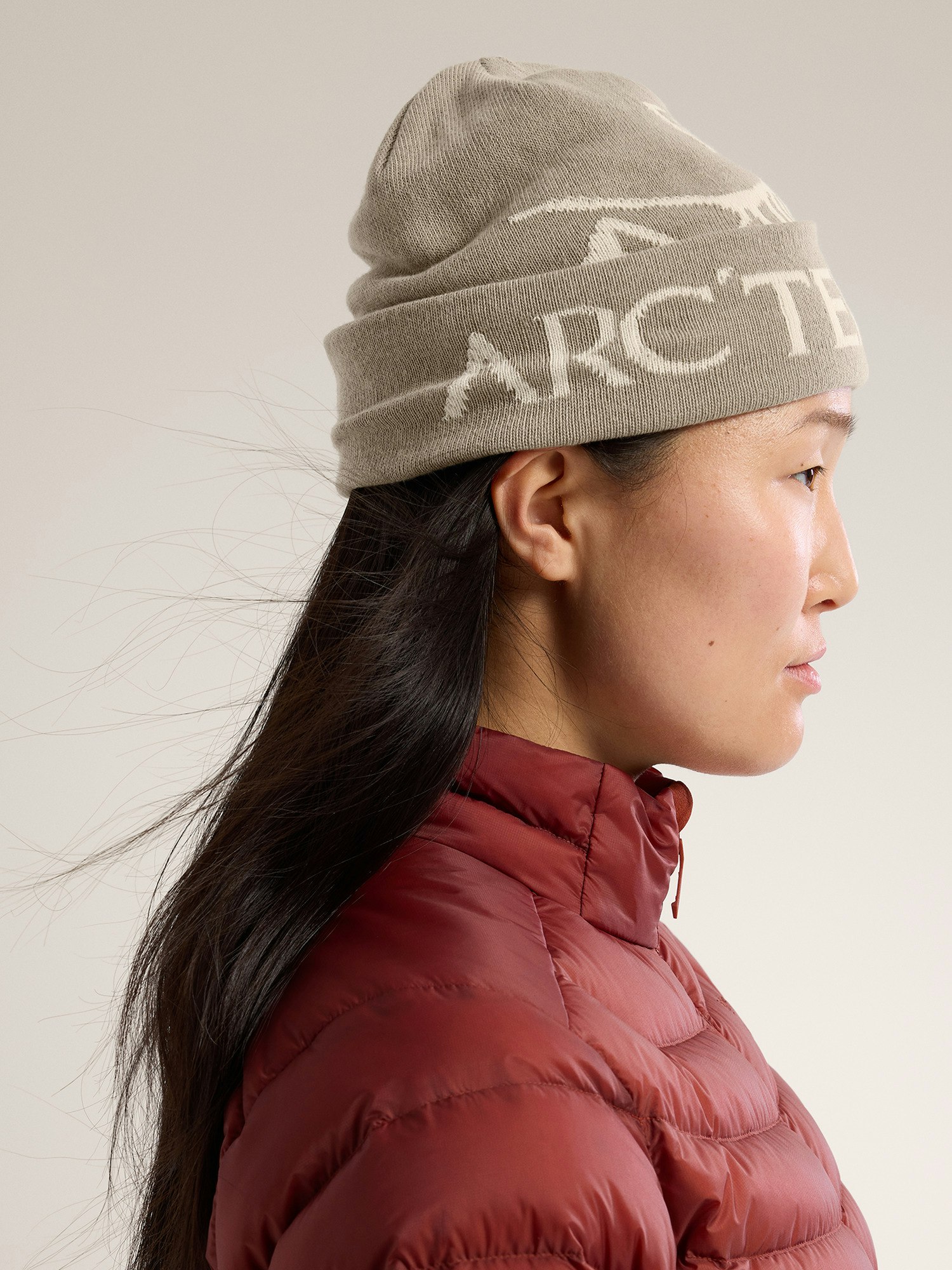 closeup side profile view of a woman wearing a grey arc'teryx beanie with an offwhite logo. she is also wearing a maroon puffer jacket. the background is a light beige.
