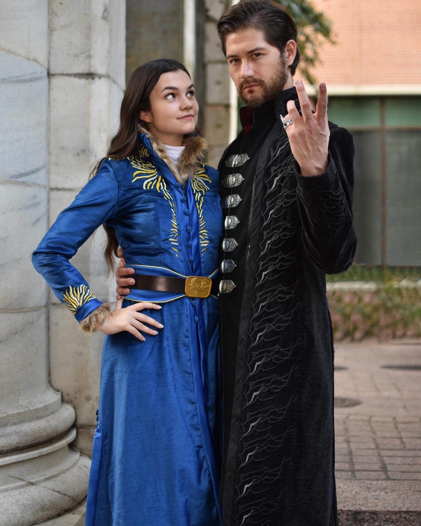 the image shows a couple in elaborate fantasystyle costumes. the woman wears a long, flowing blue dress with gold embroidery and a fur collar. the man wears a long black coat with silver detailing and is wearing a ring. they are standing close together, the man's arm around the woman's waist, posing outdoors near classical stone columns. the overall style and attire suggest a medieval or renaissanceinspired setting, possibly cosplay of specific characters.  the image has a romantic and slightly dramatic feel.
