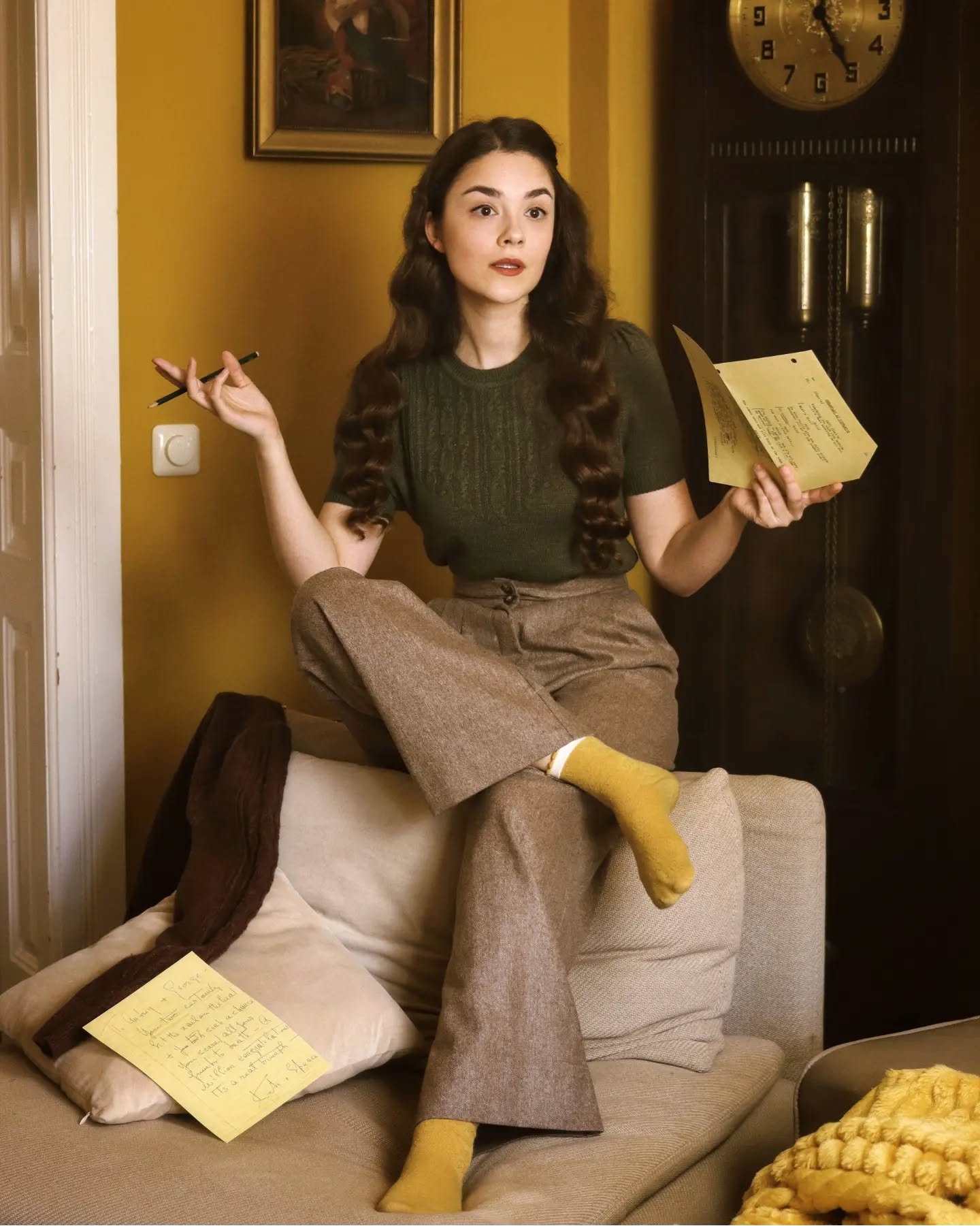 a woman in retro 1940s or 1950s style clothing sits on a sofa, holding a letter and a pencil. she is wearing a brown highwaisted trouser suit, mustard yellow socks, and has long brown hair. the setting is a cozy room with antique furniture, including a grandfather clock. the overall mood is calm and sophisticated.