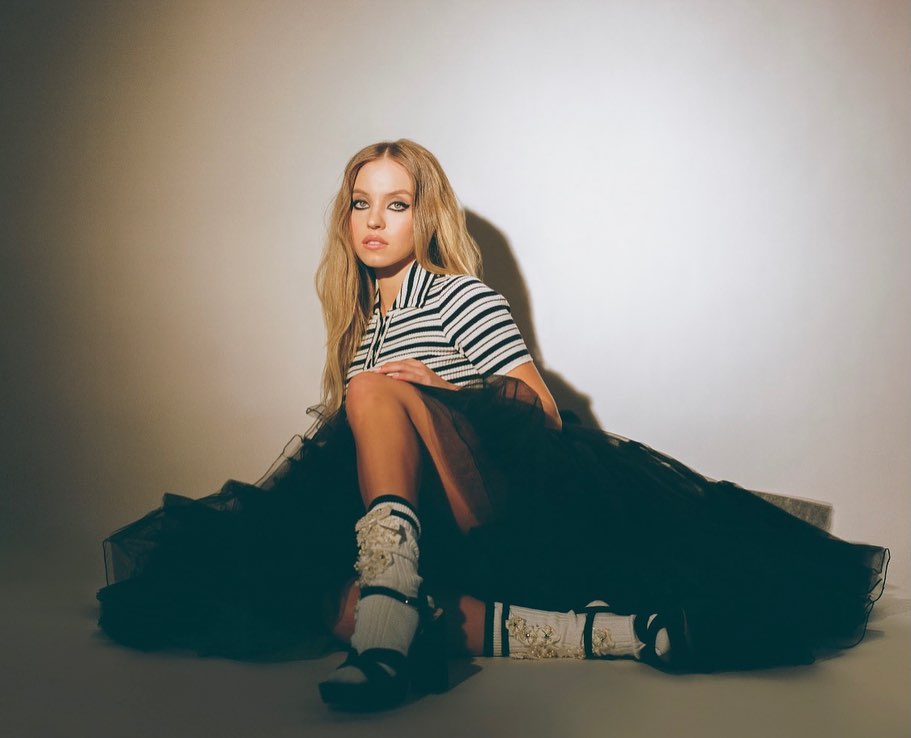 a young blonde woman is sitting on a studio floor, wearing a black tulle skirt and a striped shirt. she is wearing decorative socks and sandals. the image is a fashion photoshoot. the background is a lightcolored wall, with shadows cast on the floor.  the overall feel is elegant and stylish.