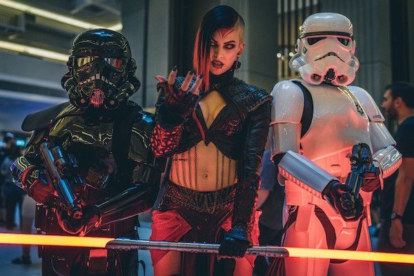 three individuals dressed in elaborate star wars costumes pose for a photo. the person in the middle is dressed in a dark, revealing outfit, while the figures flanking her are dressed as a death trooper (left) and a stormtrooper (right). they are holding weapons and a glowing lightsaber. the setting appears to be indoors, possibly at a convention center, and the lighting is primarily artificial with a neon glow.