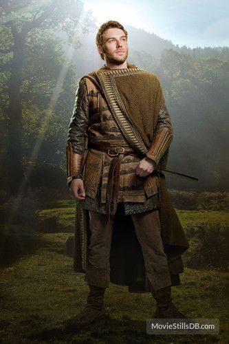a full shot of a man dressed in medieval garb, standing in an outdoor setting. he is wearing armor, a costume, and a cloak. the background features trees and a natural landscape.