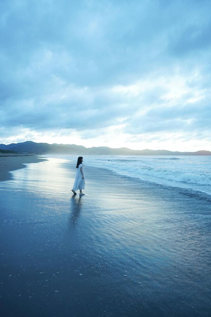 a full shot of a woman in a white dress walking on a beach. the sea is on the right, the sky is overcast with clouds, and mountains are in the background. the sand is wet and reflecting the sky.
