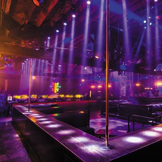 this is a wide view of the interior of a nightclub. the space is dark and lit with colorful stage lights in purple, pink, and blue hues. there's a bar in the background and what appears to be a stage with a rainbow flag. the foreground features a long bar with bar stools.
