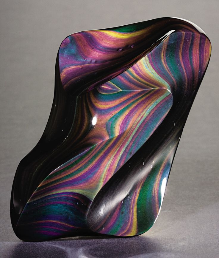 a polished piece of rainbow obsidian is shown. it has a black base color with swirls of iridescent colors including purple, blue, green, yellow, and orange. the stone is sculpted into an abstract shape and appears smooth. it is sitting on a gray surface.