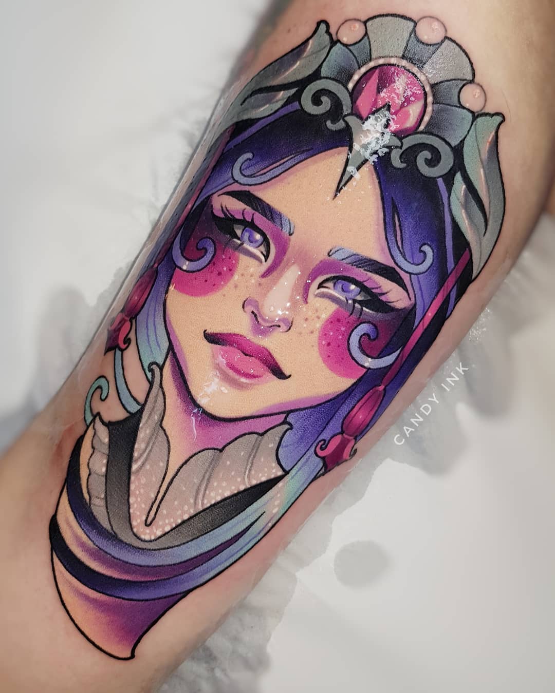 a closeup photograph showcasing a neotraditional style tattoo of a woman's face and upper torso on someone's inner arm. the woman in the tattoo has purple hair, pale skin, and rosy cheeks. she wears an elaborate headdress with a pink gem. the tattoo is done in a vibrant color palette with bold linework. the tattoo artist's signature "candy ink" is visible in a light script below the tattoo.