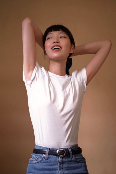 a young asian woman with short black hair is smiling while looking at the camera. she is wearing a white tshirt and jeans, and is standing in a studio. she is looking at the camera and smiling.