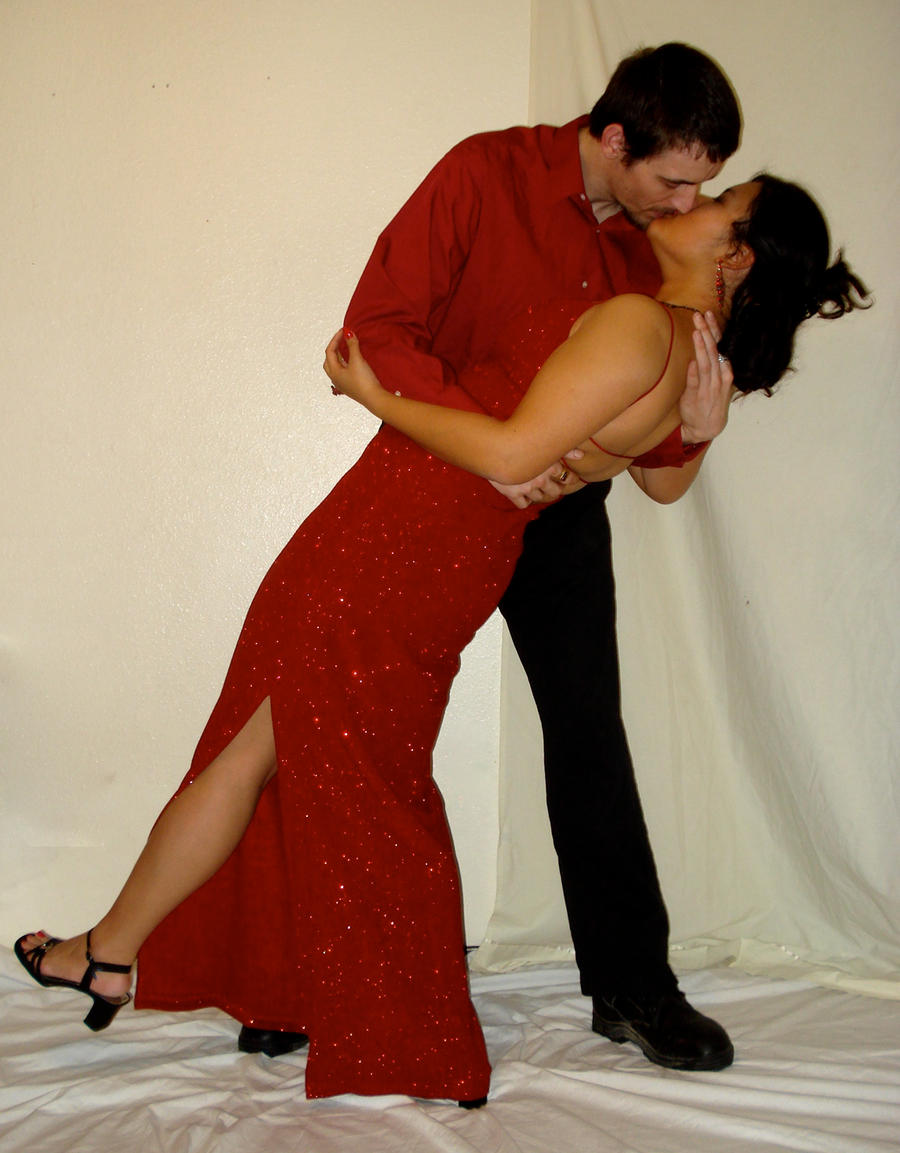 the image shows a couple passionately kissing. the woman is wearing a long, red, glittering gown with a high slit, while the man is dressed in a red shirt and black trousers.  they are embraced in a close dancelike pose, suggesting intimacy and romance. the setting appears to be indoors, against a simple white background. the overall mood is one of passionate love and elegance.