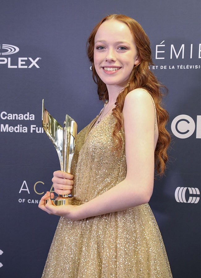 a young redhaired actress in a gold dress smiles while holding a canadian screen award trophy at an awards ceremony.  the backdrop features logos for the canadian screen awards, including sponsors like telefilm canada and the canada media fund.  the overall impression is one of celebration and success.