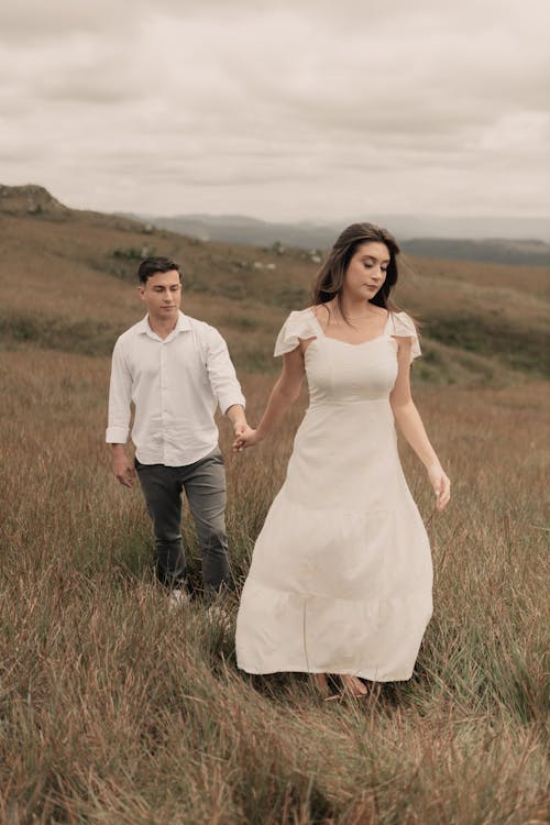 a couple, a man and a woman, are walking handinhand through a tall grass field. the woman is wearing a long, white dress and the man is wearing a white longsleeved shirt and grey trousers.  they are walking towards the viewer. the setting is outdoors, with a landscape of rolling hills and a cloudy sky in the background. the overall mood is romantic and serene.