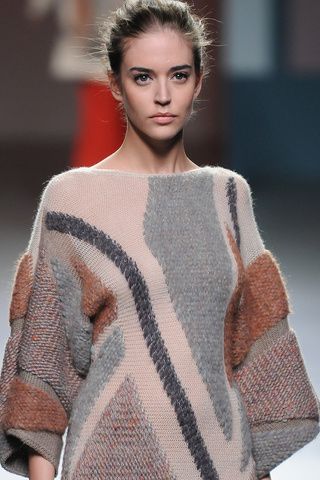 a young woman with short brown hair is wearing a gray, brown and pink knitted sweater. she is looking at the camera, and her hair is slightly parted on one side. the sweater has a geometric design. the background is blurred, but the focus is on the model and her outfit.
