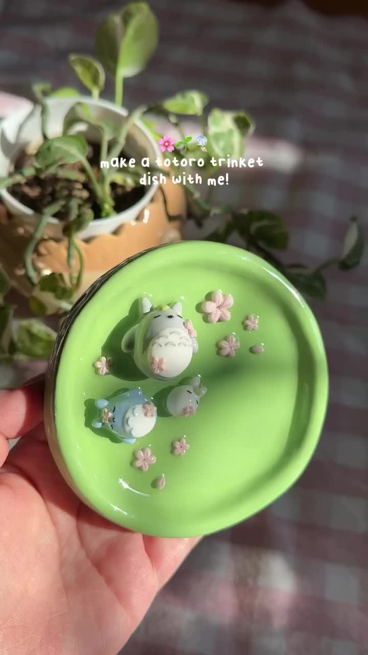 a handmade totoro trinket dish with totoro and chibi totoro figurines along with pink flower decorations is held in a hand. a plant in a decorative pot can be seen in the background.