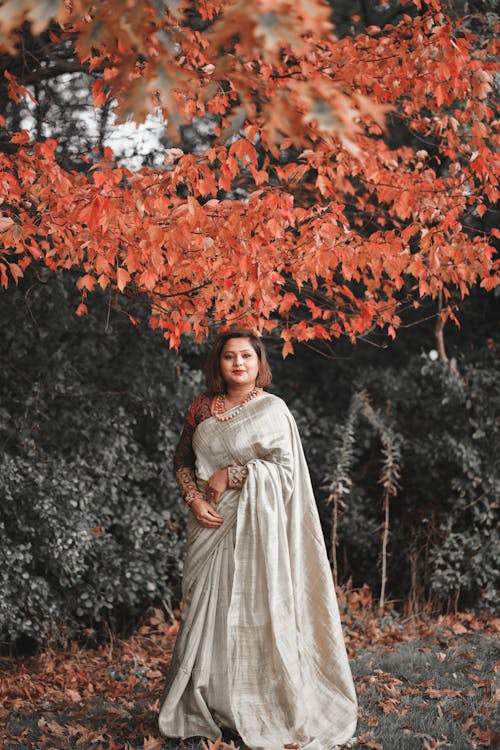a woman in a flowing light grey silk saree stands under a tree with vibrant orangered autumn leaves. she is looking calmly at the camera. the background is slightly desaturated, emphasizing the colors of the saree and leaves.  the overall mood is serene and elegant.