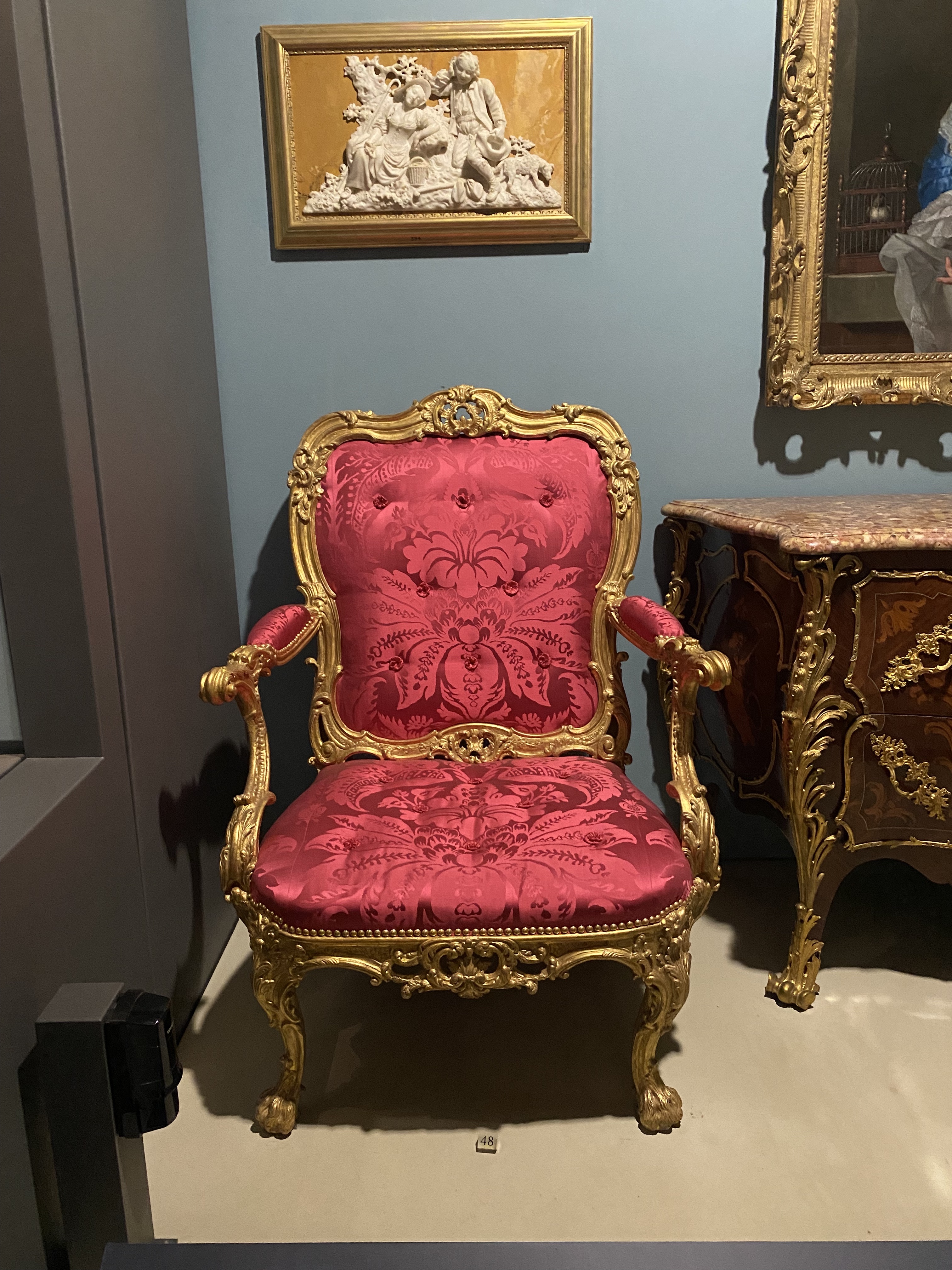 the image shows a luxurious red and gold ornate chair in a museum setting, likely an example of rococo or louis xv style furniture. the chair is surrounded by other pieces of art and furniture, including a framed sculpture and a gilded chest of drawers. the overall impression is one of opulence and historical significance.
