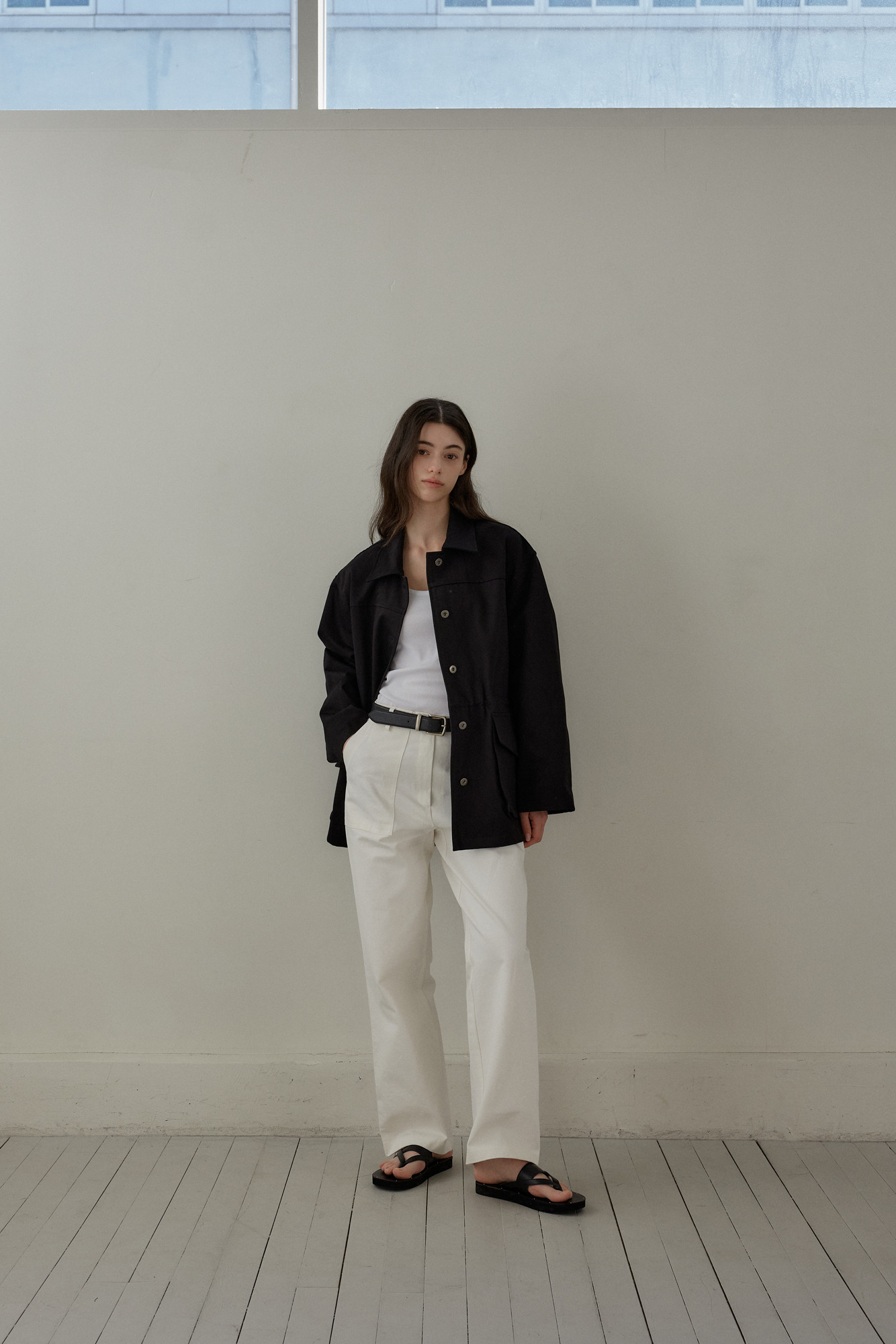 a full shot of a woman standing indoors. she wears a white tank top, a black jacket, white pants and black sandals. she has a black belt on her pants. her hair is dark brown and long. she is posing in front of a light grey wall. there is a window in the upper part of the frame. the woman is dressed casually.