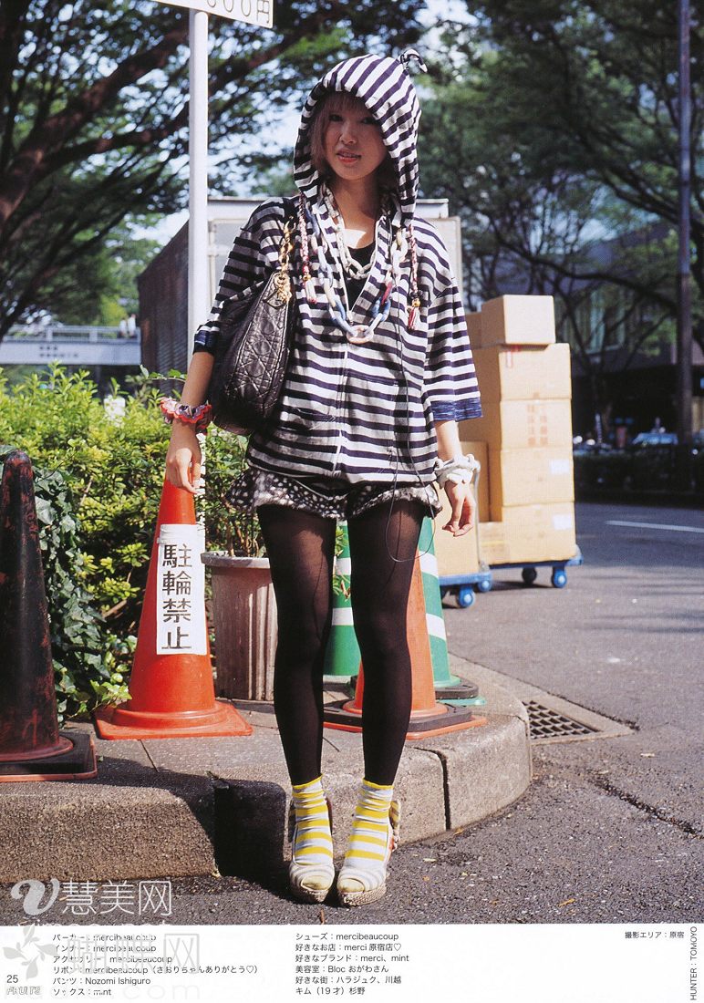 the image shows a young woman posing on a sidewalk, her back to a row of shops or houses. she is wearing a black and white striped hooded top over a black dress, black tights and yellow platform sandals. she is carrying a large black shoulder bag. the overall style of her clothing suggests a casual, streetstyle aesthetic. the background is slightly out of focus, drawing attention to the woman in the foreground. the lighting suggests it is a sunny day, with the sunlight illuminating the woman from the back and creating a slight halo effect around her hair.