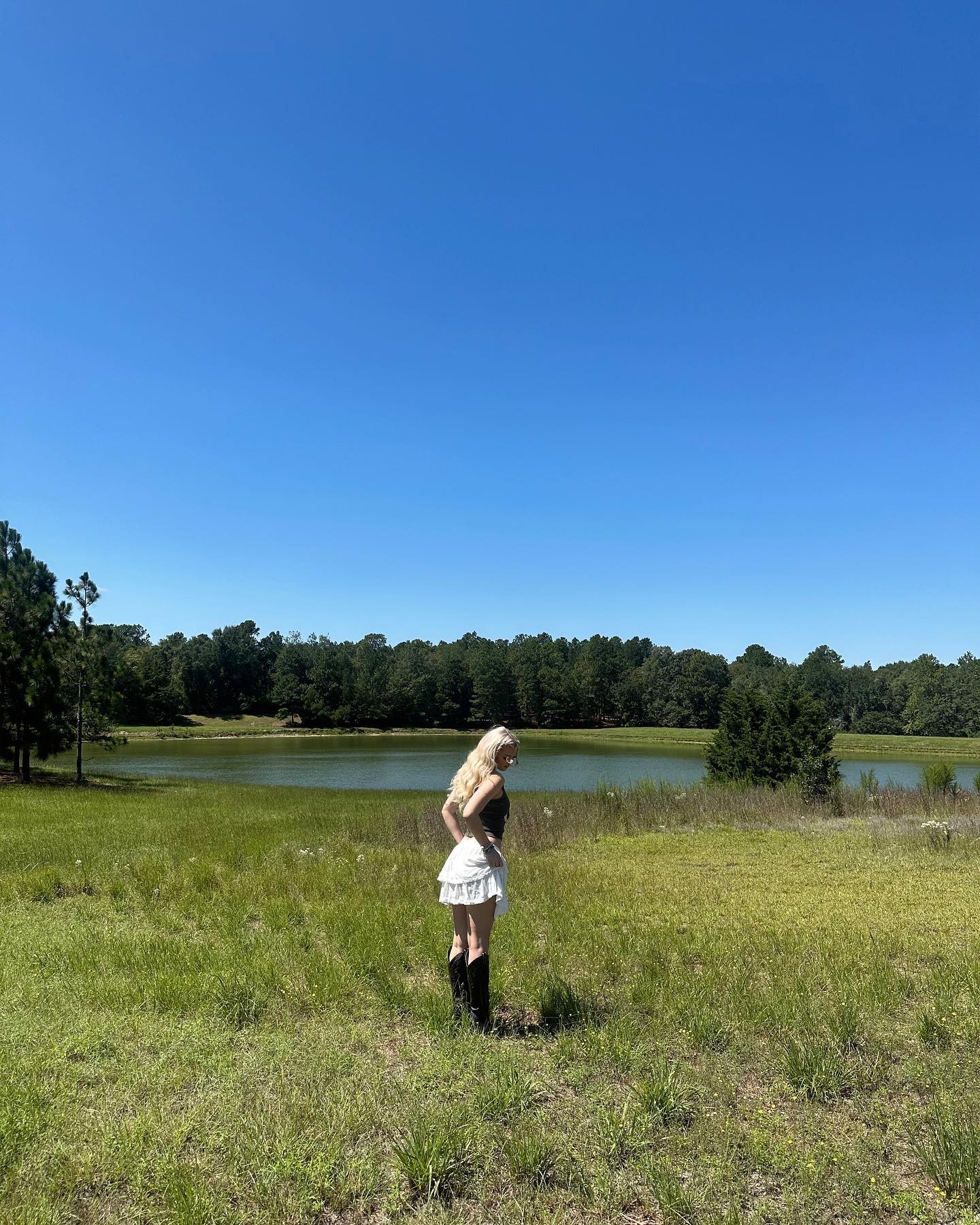 a young woman with blonde hair stands in a grassy field, wearing a white skirt, black top, and boots. behind her is a lake surrounded by trees, under a clear blue sky. the setting appears to be a rural landscape during the day.