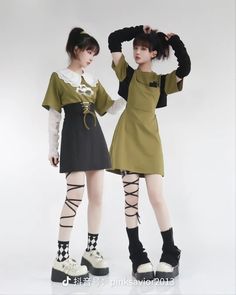 two young women pose sidebyside against a minimalist offwhite backdrop. they showcase distinct yet complementary outfits reflecting a blend of soft grunge and ulzzang styles. the woman on the left sports a white collared top layered beneath a black pinafore dress, her look finished with platform shoes, white socks with black accents, and a halfup hairstyle. on the right, her friend wears an olive green dress featuring a unique strap detail, paired with black platform shoes, black socks, arm warmers, and a black beret, creating an effortlessly chic ensemble. the image captures their confident poses and highlights the details of their trendy attire.