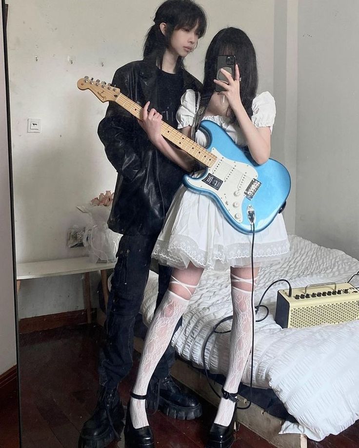 he image shows two young asian women, one dressed in a black leather jacket and the other in a white lace dress, playing an electric guitar together in what appears to be a bedroom. the guitar is light blue. the woman in the dress is wearing white lace thighhigh socks. the scene is presented in an aesthetic, alternative style.  a mirror reflects a portion of the room.