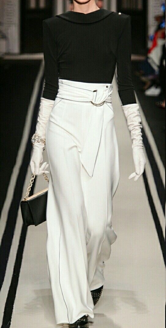 a fashion model is walking down the runway wearing white wideleg pants, a black top, and white gloves. she has a black clutch bag with a silver chain attached to it. she is wearing a belt with a large silver buckle. she is styled in a glamorous and sophisticated look.