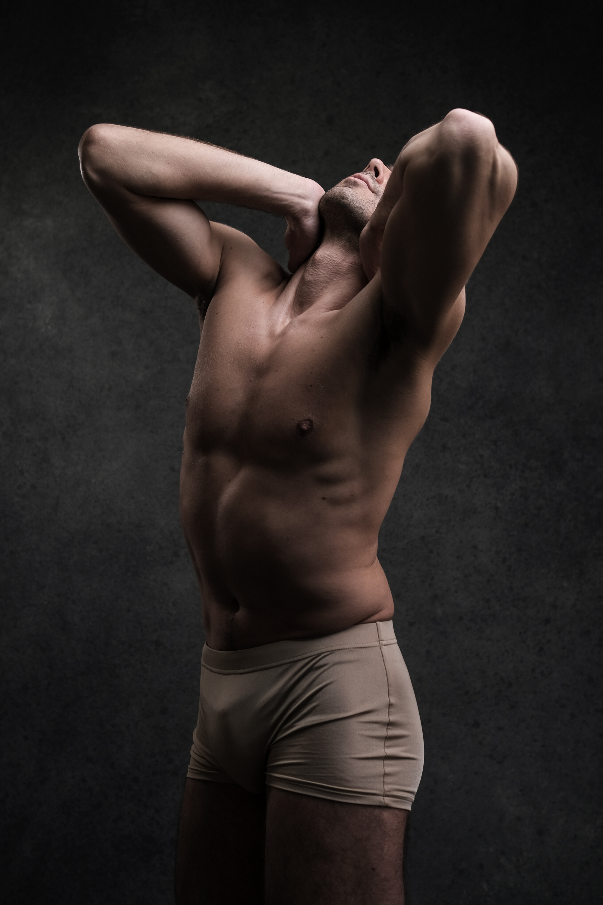 a muscular man in beige underwear, illuminated by dramatic lighting, poses against a dark background, his arms raised and hands clasped behind his head, highlighting his physique and strength.